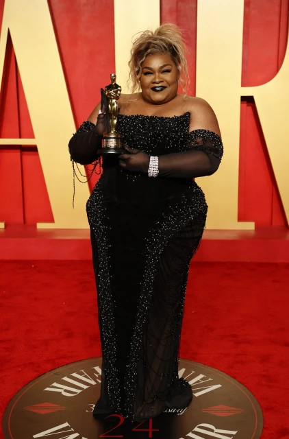 Woman in black gown holds Oscar award on red carpet. Blonde hair, dark makeup, statement bracelet. Red and gold backdrop, full shot.