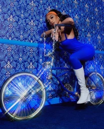 Woman in blue jumpsuit posing on a custom blue bicycle with glittery chrome handlebars against blue patterned wallpaper