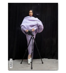 Woman in oversized lavender ribbed sweater standing behind camera on tripod against black curtain backdrop