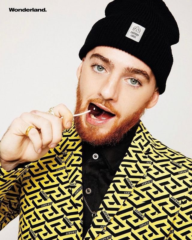 Close-up of an adult male in a yellow Versace blazer, wearing a black beanie and eating a lollipop against a white background.