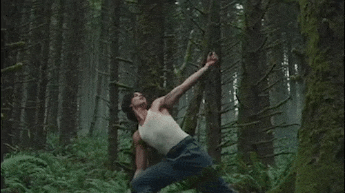 An athletic man in a white tank top and jeans dances expressively in a dense, lush forest with tall trees and abundant green foliage.