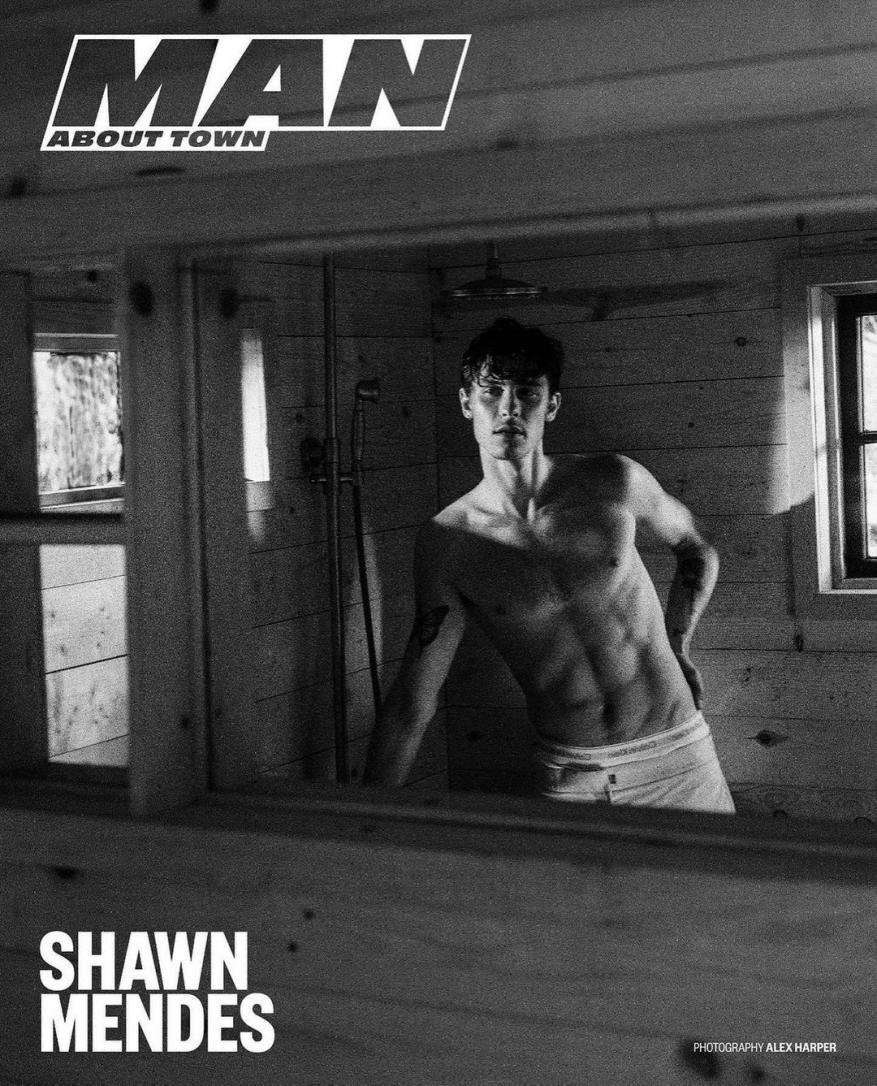 Monochrome portrait of shirtless Shawn Mendes framed by wooden shower structure, showcasing his physique and tattoo detail, in natural light.