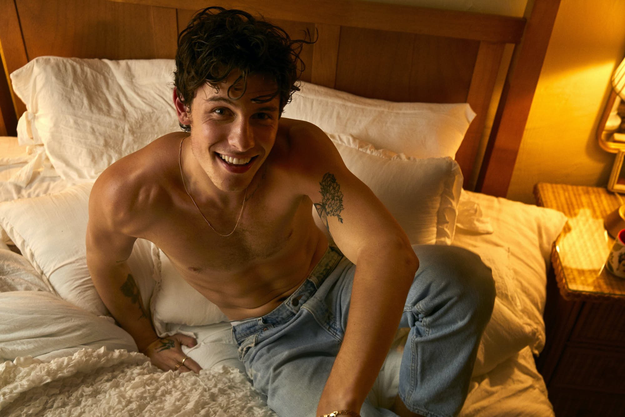 Shirtless adult male with a tattoo on his upper arm smiles while reclining on a bed with white bedding in a brightly lit room.