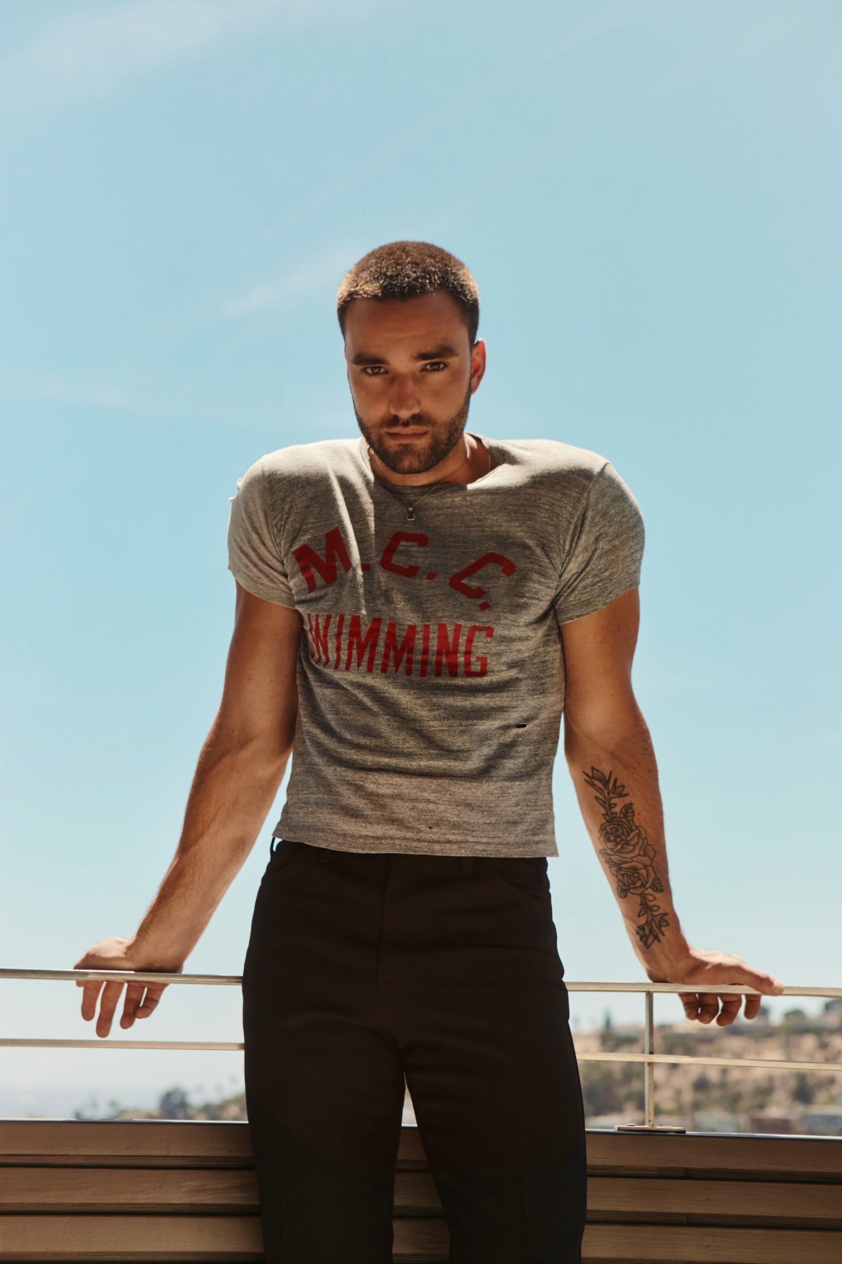 Handsome man with short hair poses in t-shirt and black jeans on balcony. Clear sky provides a bright, natural light backdrop.