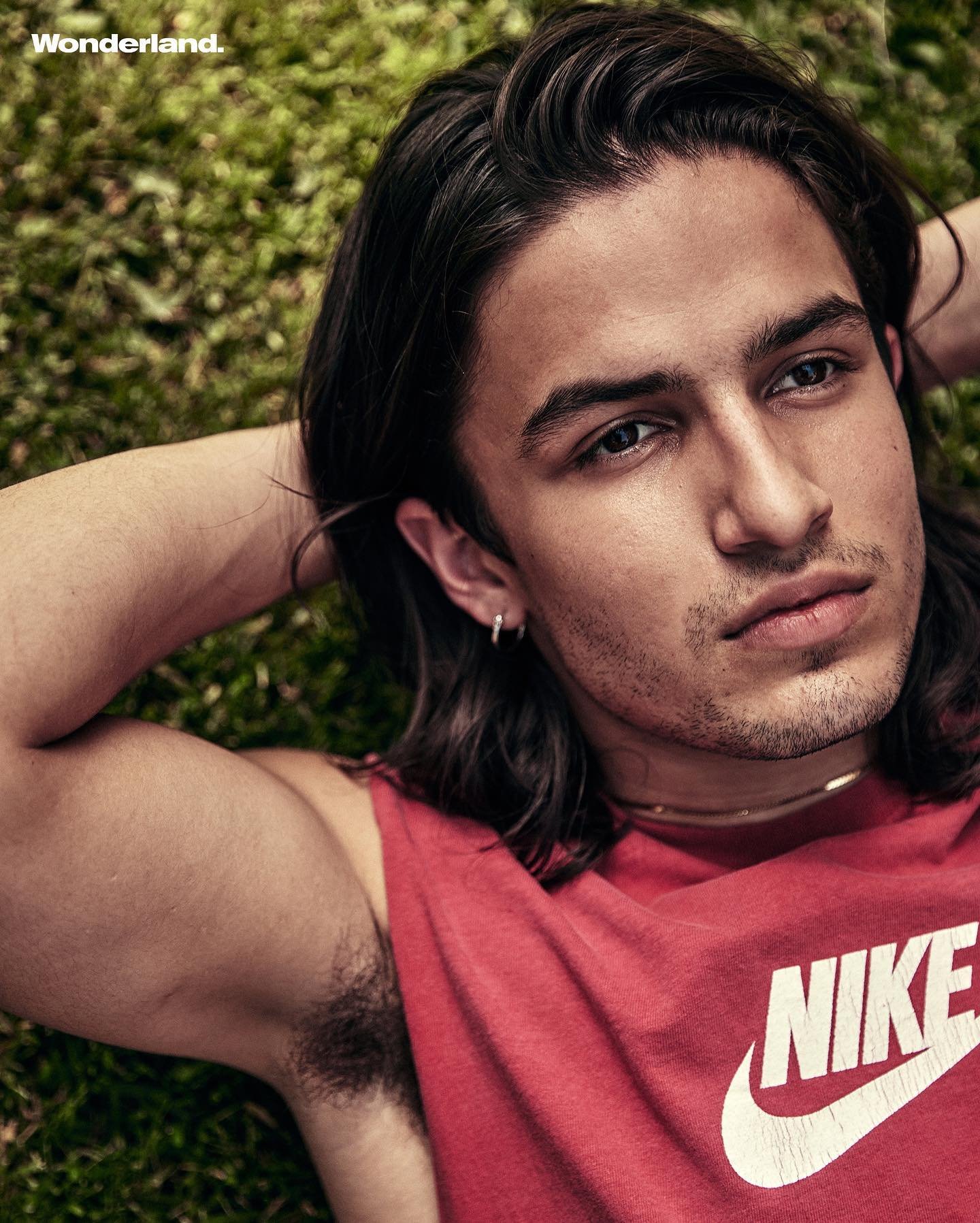 Portrait of a young adult male with long brown hair, wearing a red Nike tank top, laying on green grass, looking at the camera.