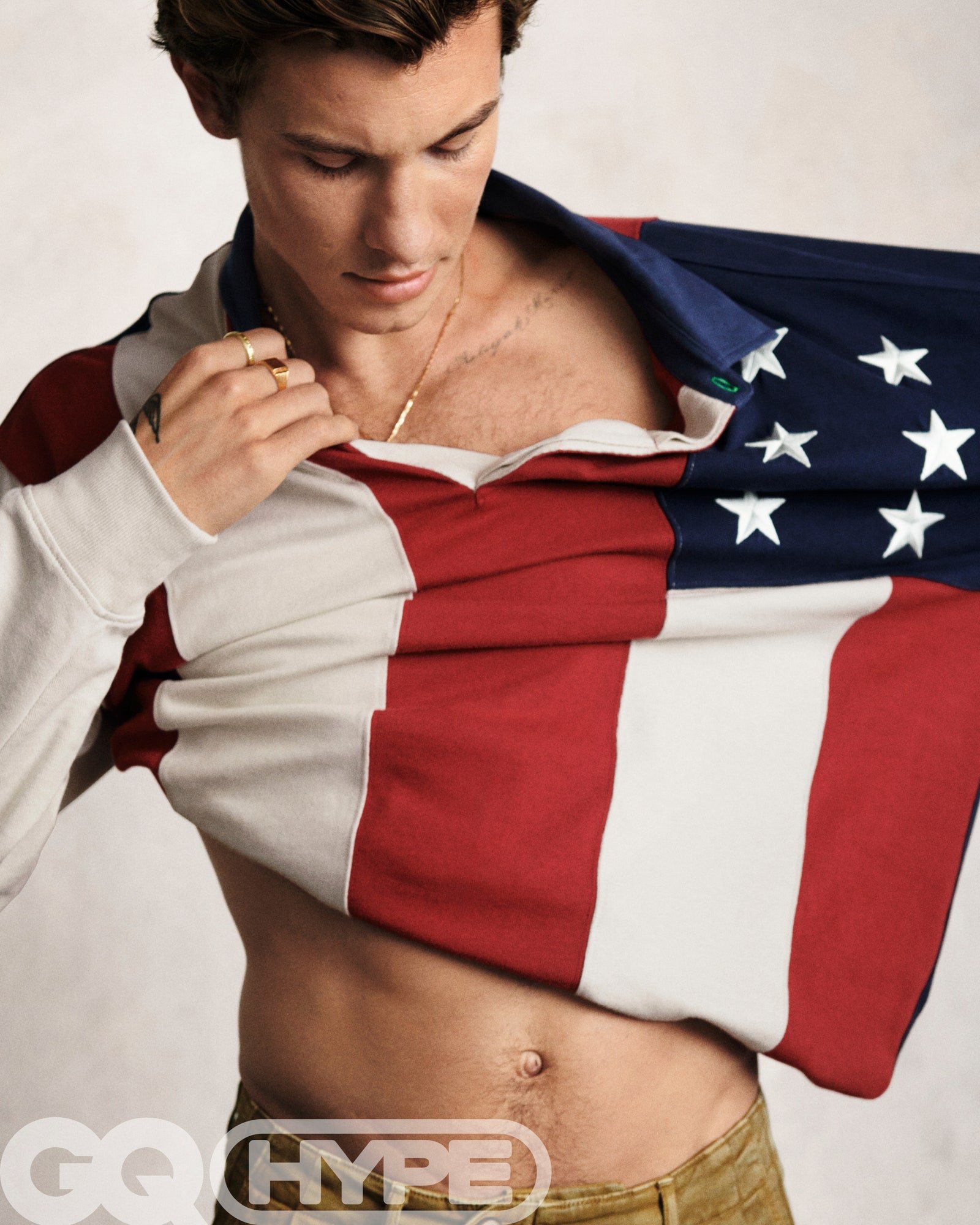 Male model, shirtless, wearing American flag-themed shirt and tan pants, poses in a close-up studio shot for a fashion editorial.