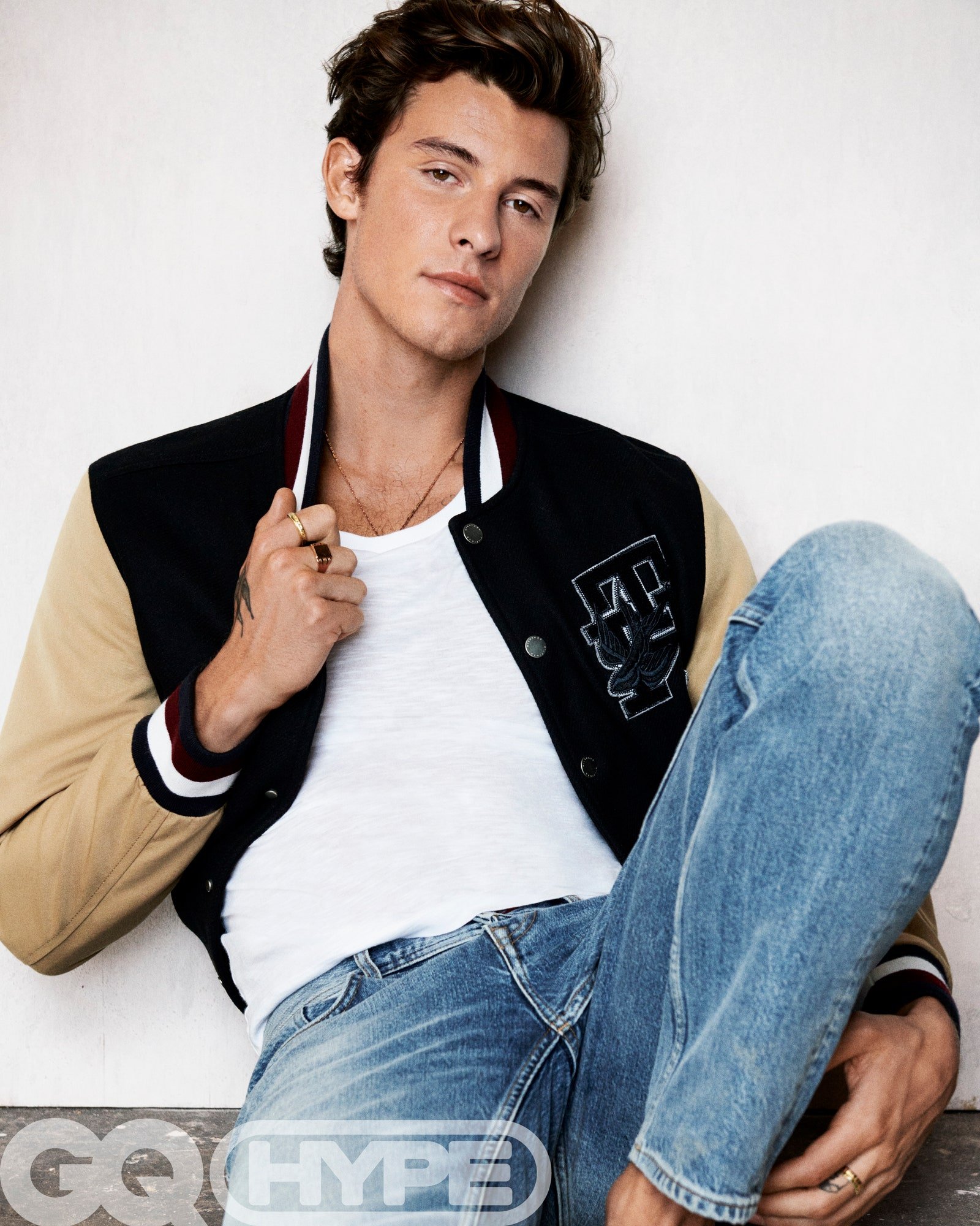 Portrait of Shawn Mendes wearing a bomber jacket, white T-shirt, and blue jeans, seated against a white wall in a photo studio setting.