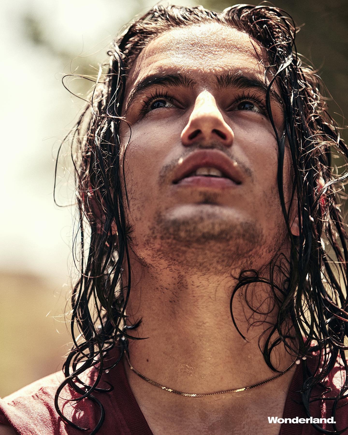 Close-up portrait of a young adult male with wet, long hair, gazing upwards with a neutral expression, wearing a gold chain necklace.