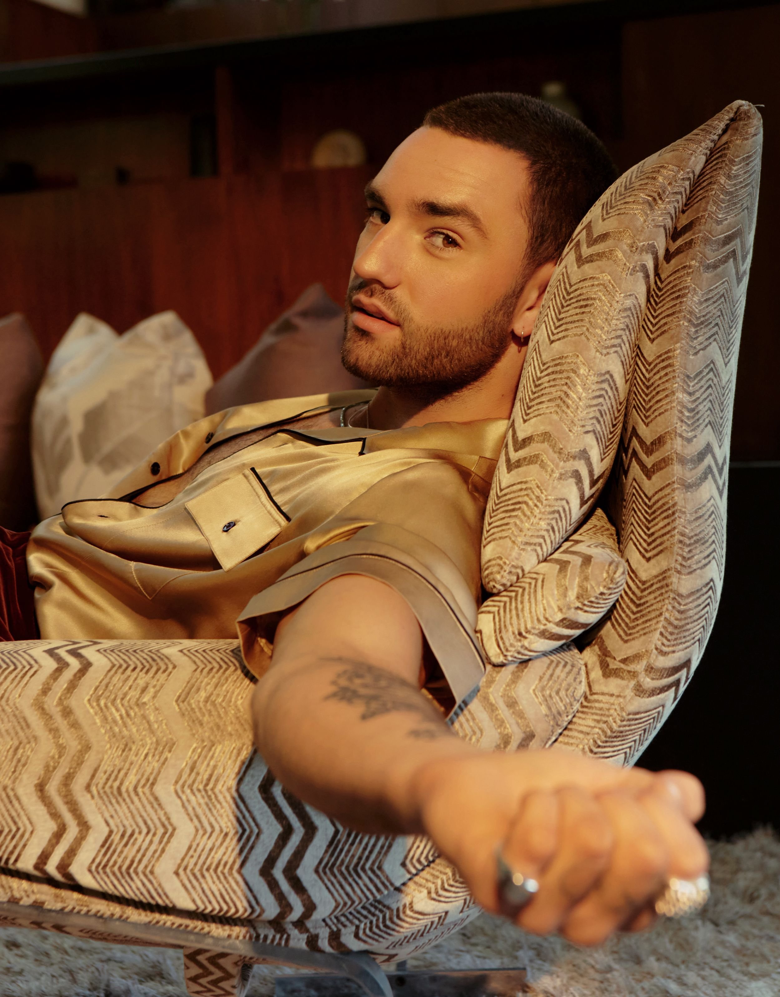 An adult male with a beard is relaxing in a patterned armchair, wearing a gold satin pajama shirt, indoors, with a dark wood background.