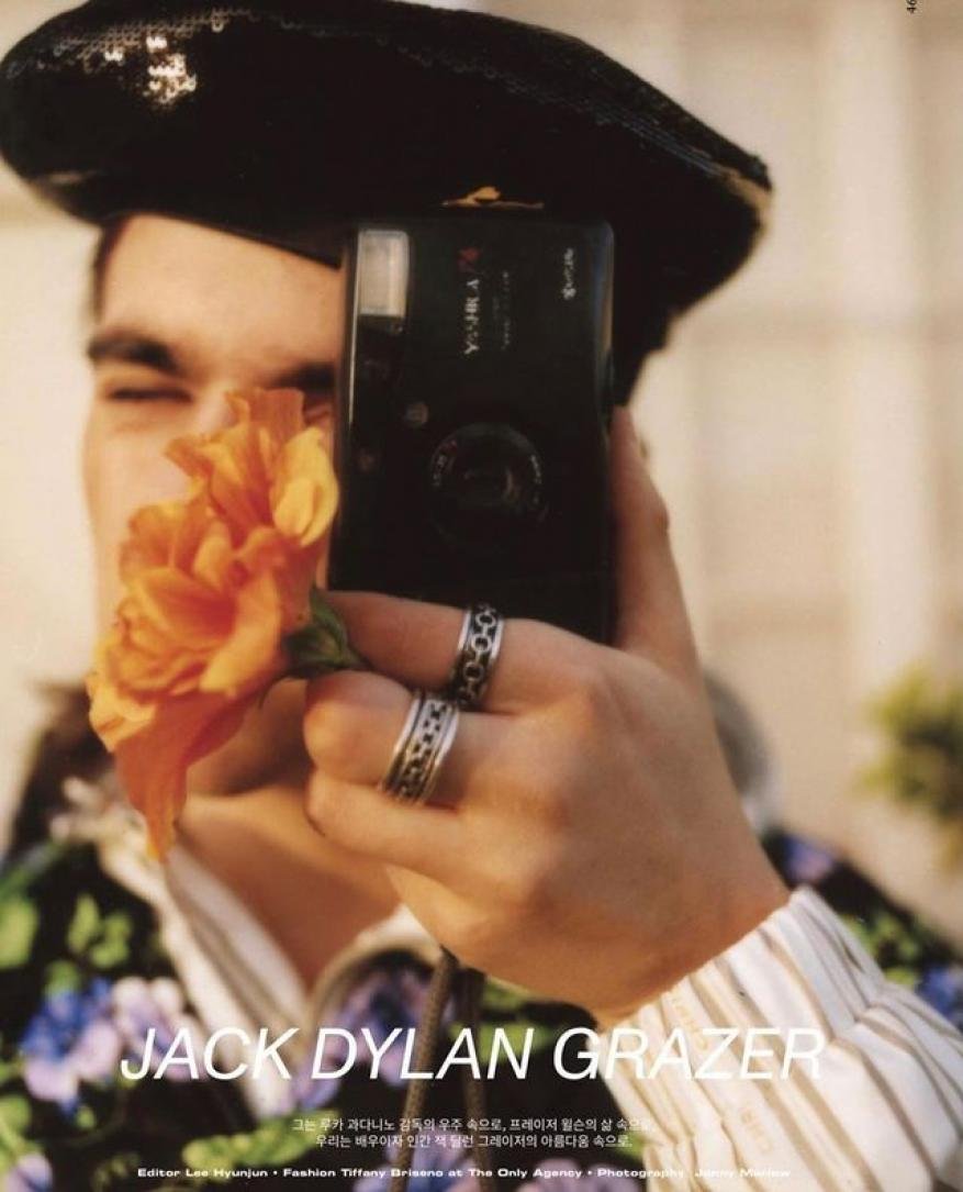 Jack Dylan Grazer holds a camera and an orange flower, wearing a black beret and a shirt with floral patterns and white stripes.