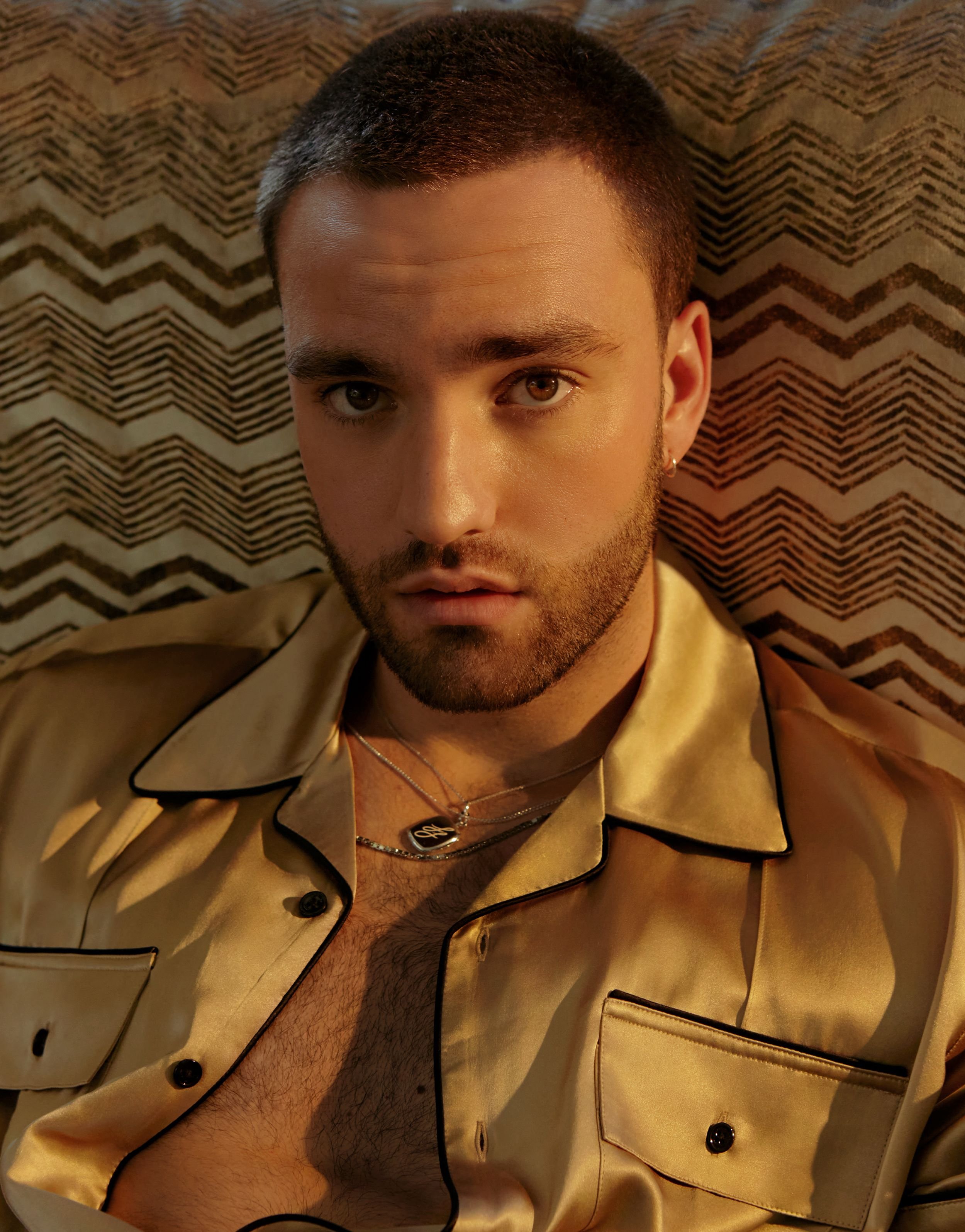 Close-up portrait of a young adult man with short brown hair and a light beard, wearing a silk shirt and necklace against a patterned backdrop.