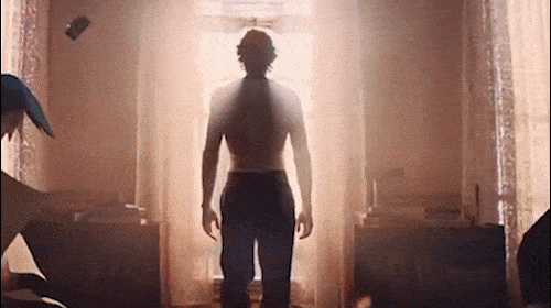 Silhouette of a shirtless man standing in a sunlit doorway, with two blurred figures in a room with furniture.