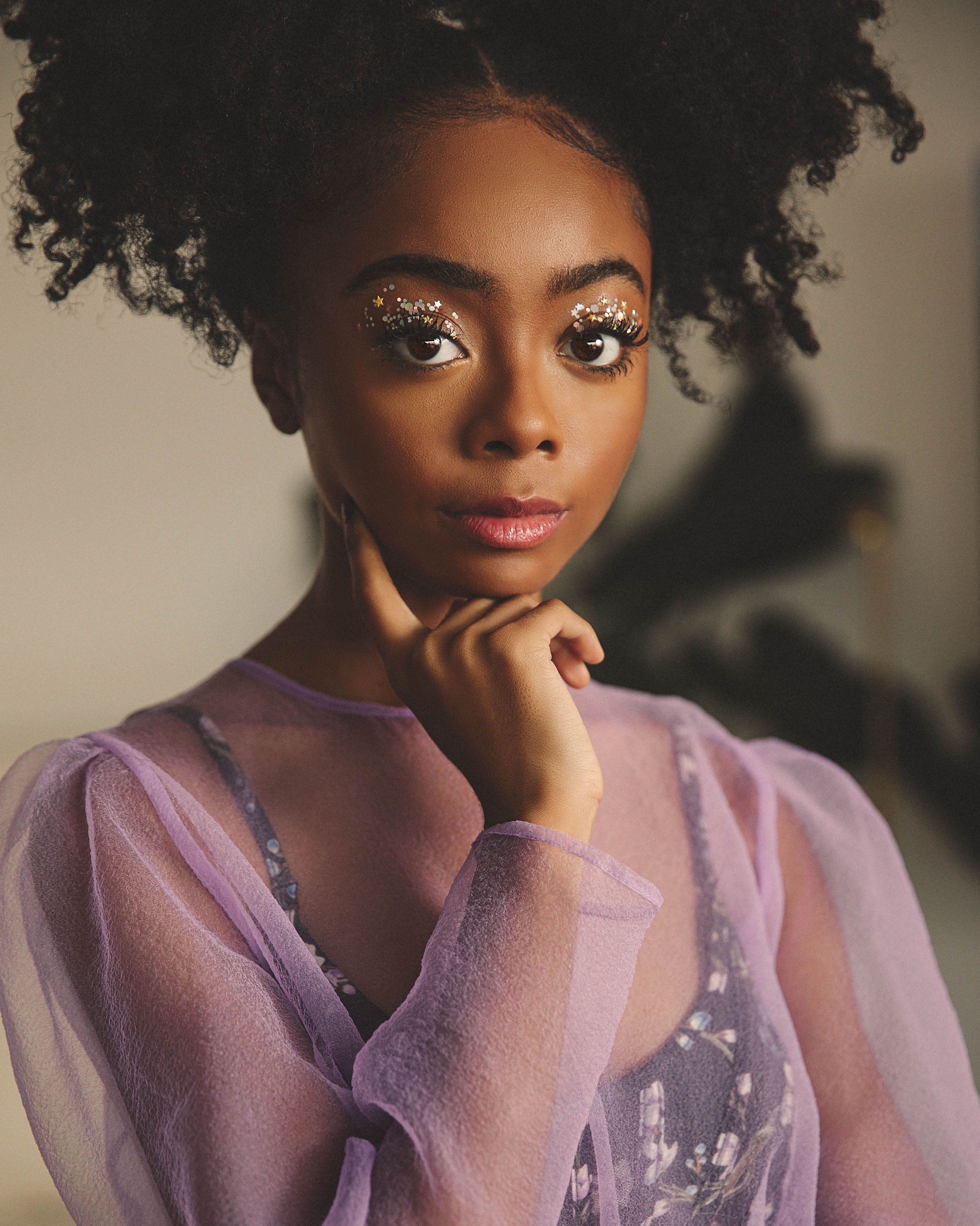 Close-up portrait of a young Black woman with glitter eye makeup, wearing a sheer purple top. She is posed indoors with soft lighting.