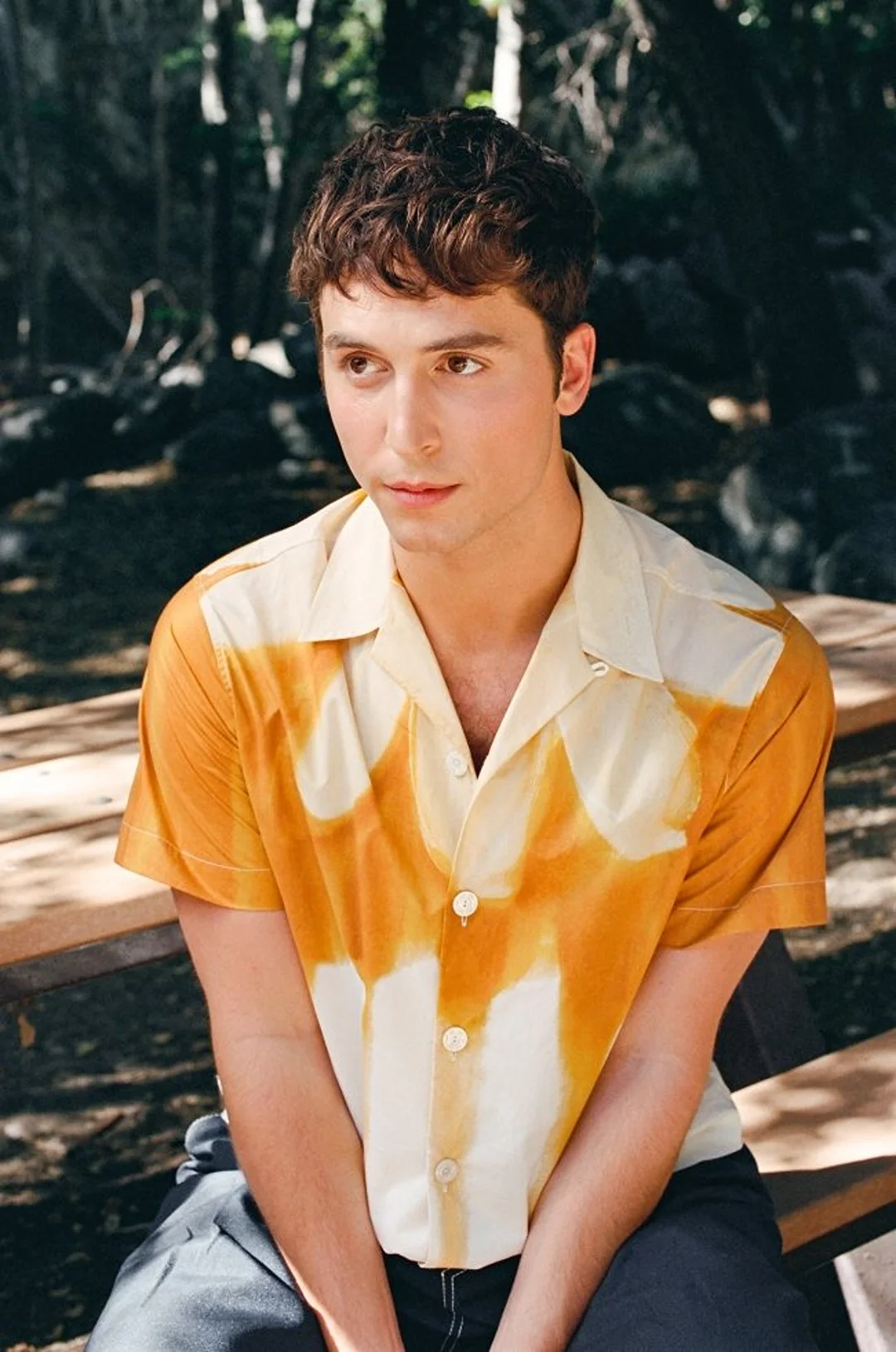 A young adult male in a patterned shirt poses outdoors in medium shot with a soft-focus background of foliage and dappled sunlight.