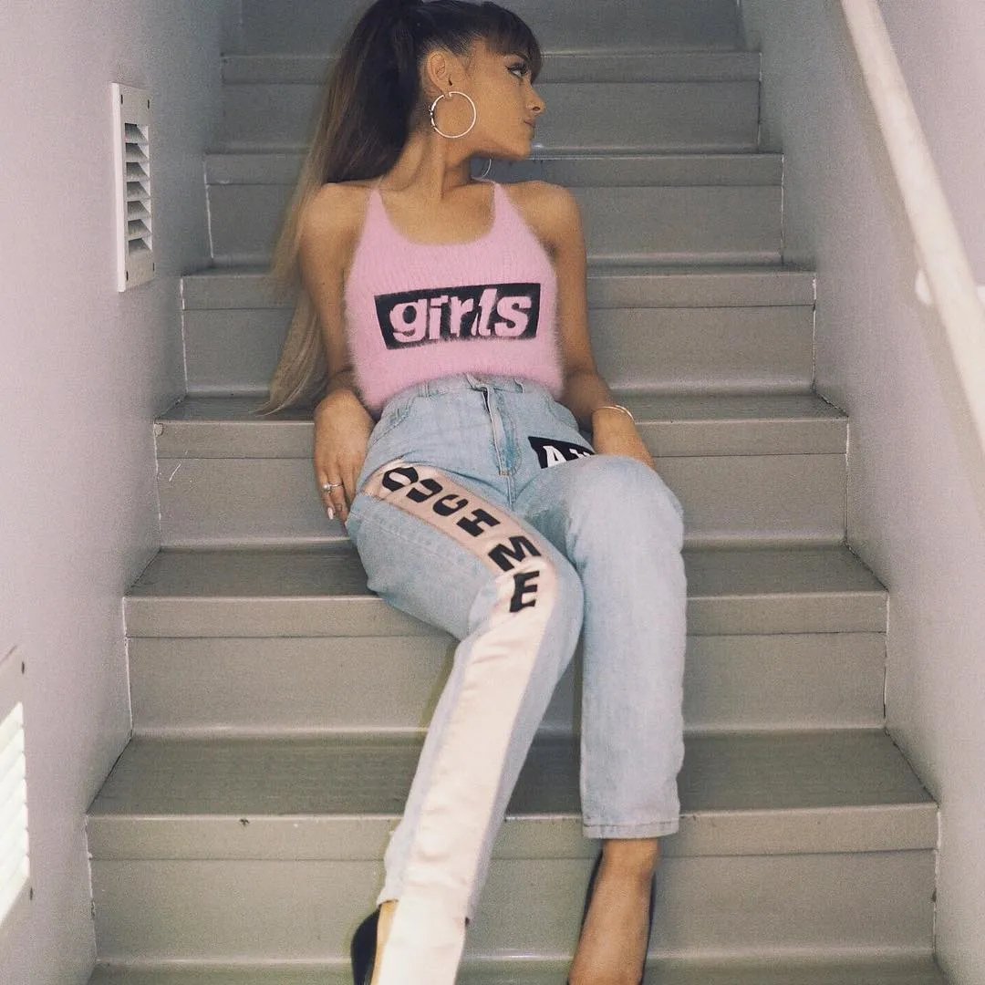 Ariana Grande sitting on grey indoor stairs wearing a pink fuzzy crop top and light blue jeans