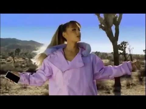 Ariana Grande wearing a lilac puffer jacket holding a smartphone in a desert landscape with Joshua trees.