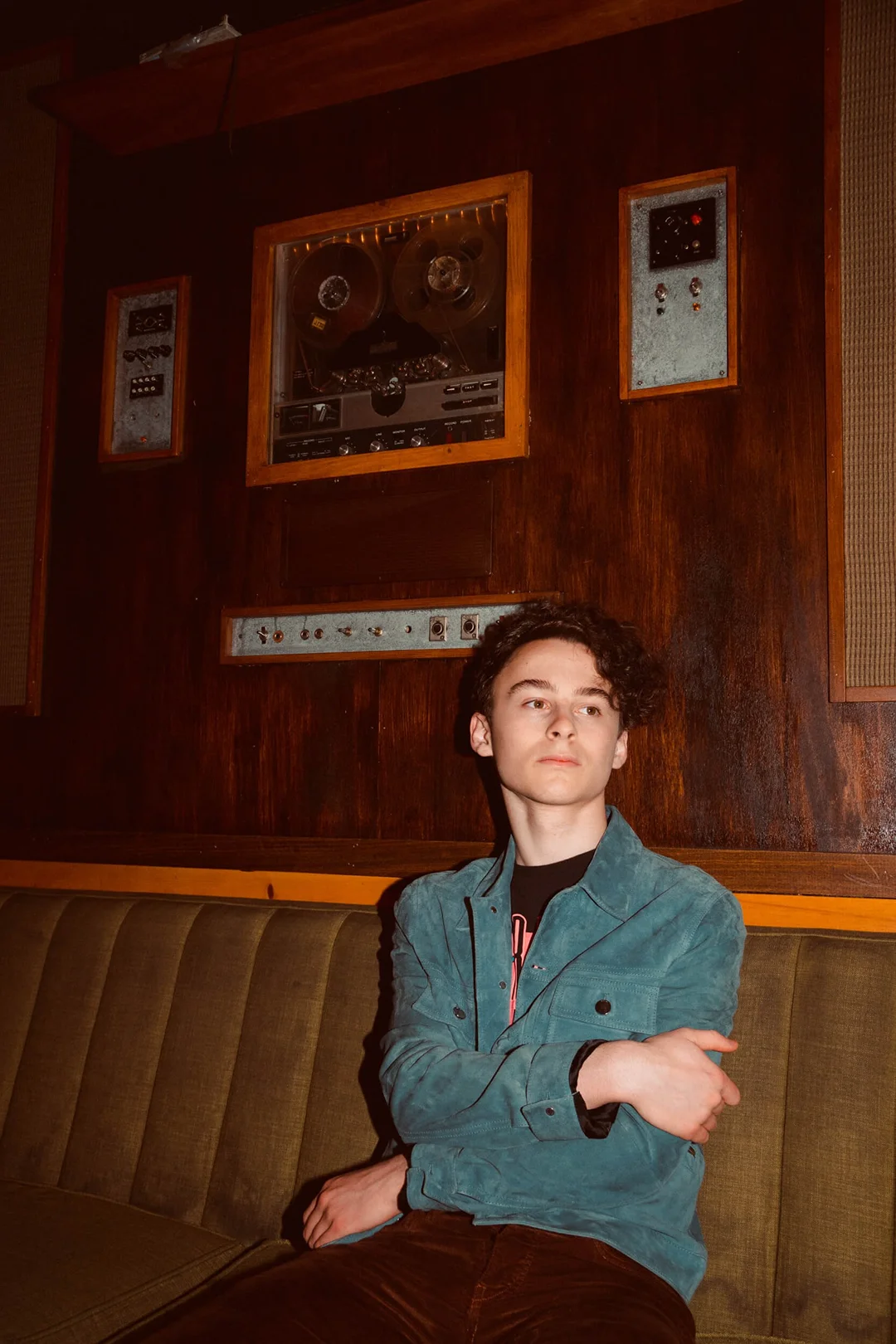 Teenage boy with curly brown hair poses on an olive-green couch in front of a dark wooden wall with vintage audio equipment.