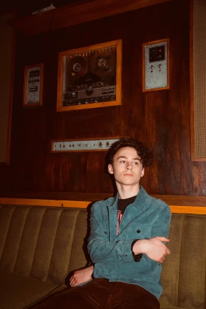 Teenage boy with curly brown hair poses on an olive-green couch in front of a dark wooden wall with vintage audio equipment.