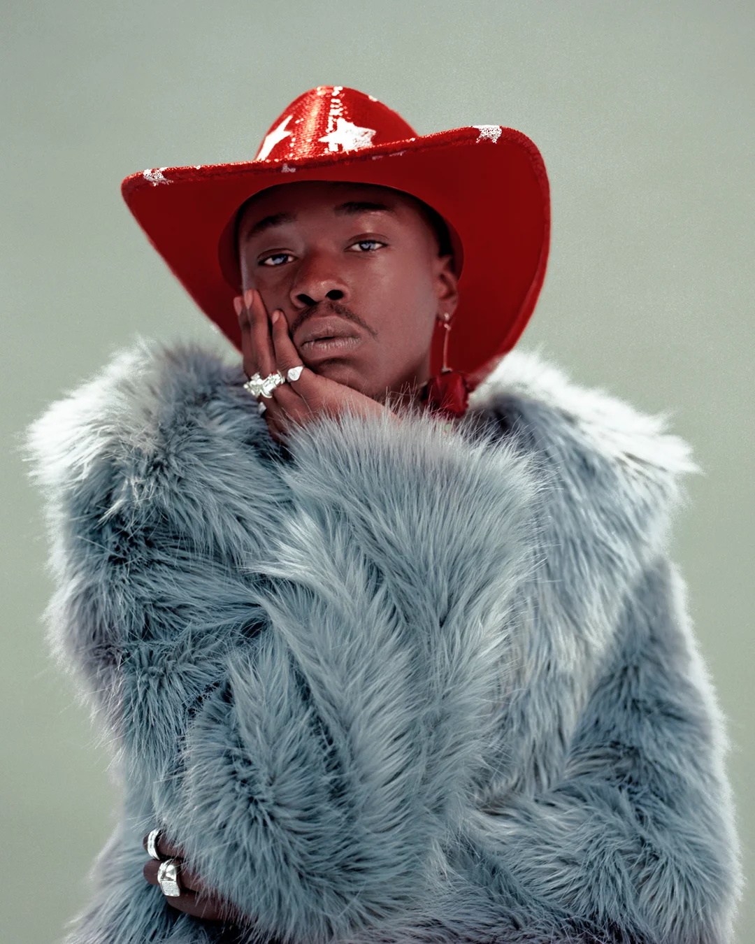 Portrait of Black adult male wearing a red cowboy hat and fur coat, hand to face, against a neutral background. Fashion, celebrity style.