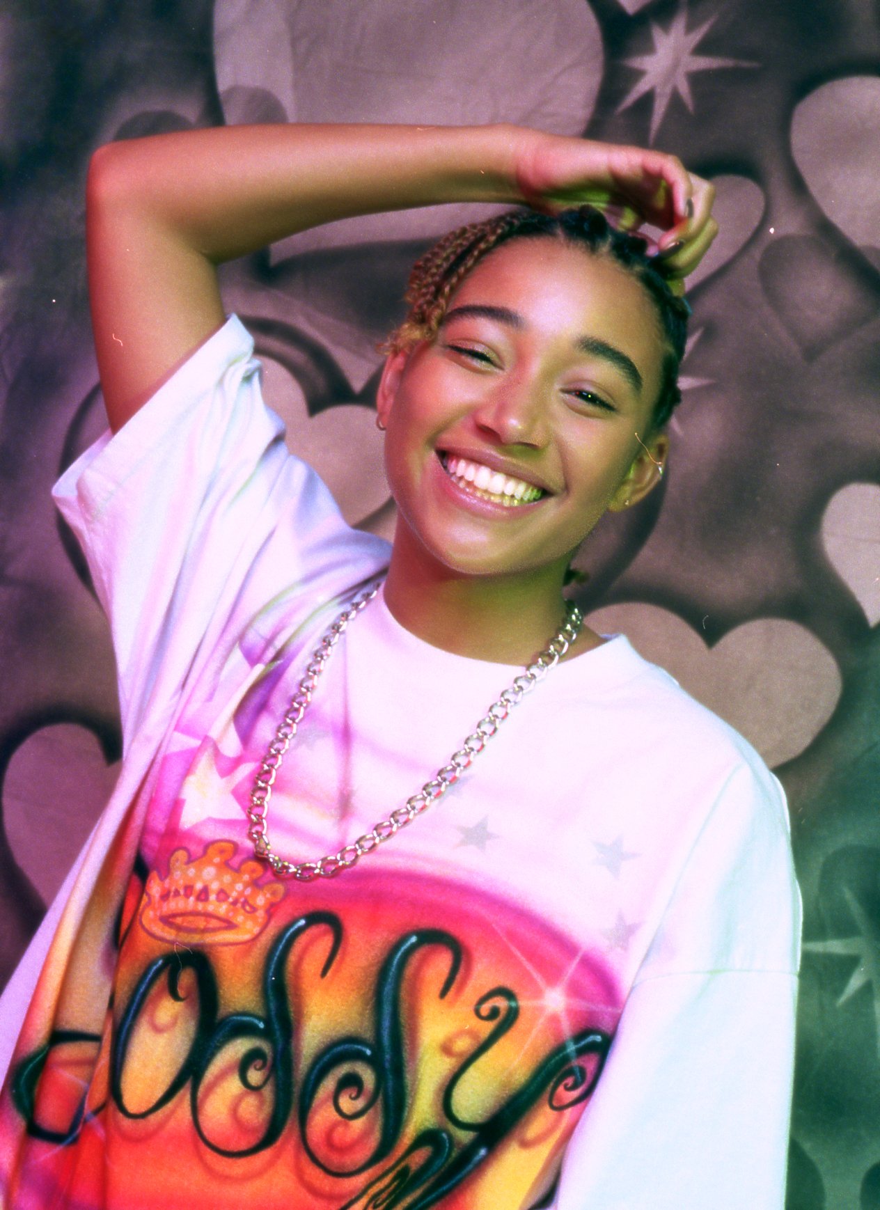 Smiling young woman in a graphic tee and silver chain posing against a colorful graffiti background with stars and heart shapes.