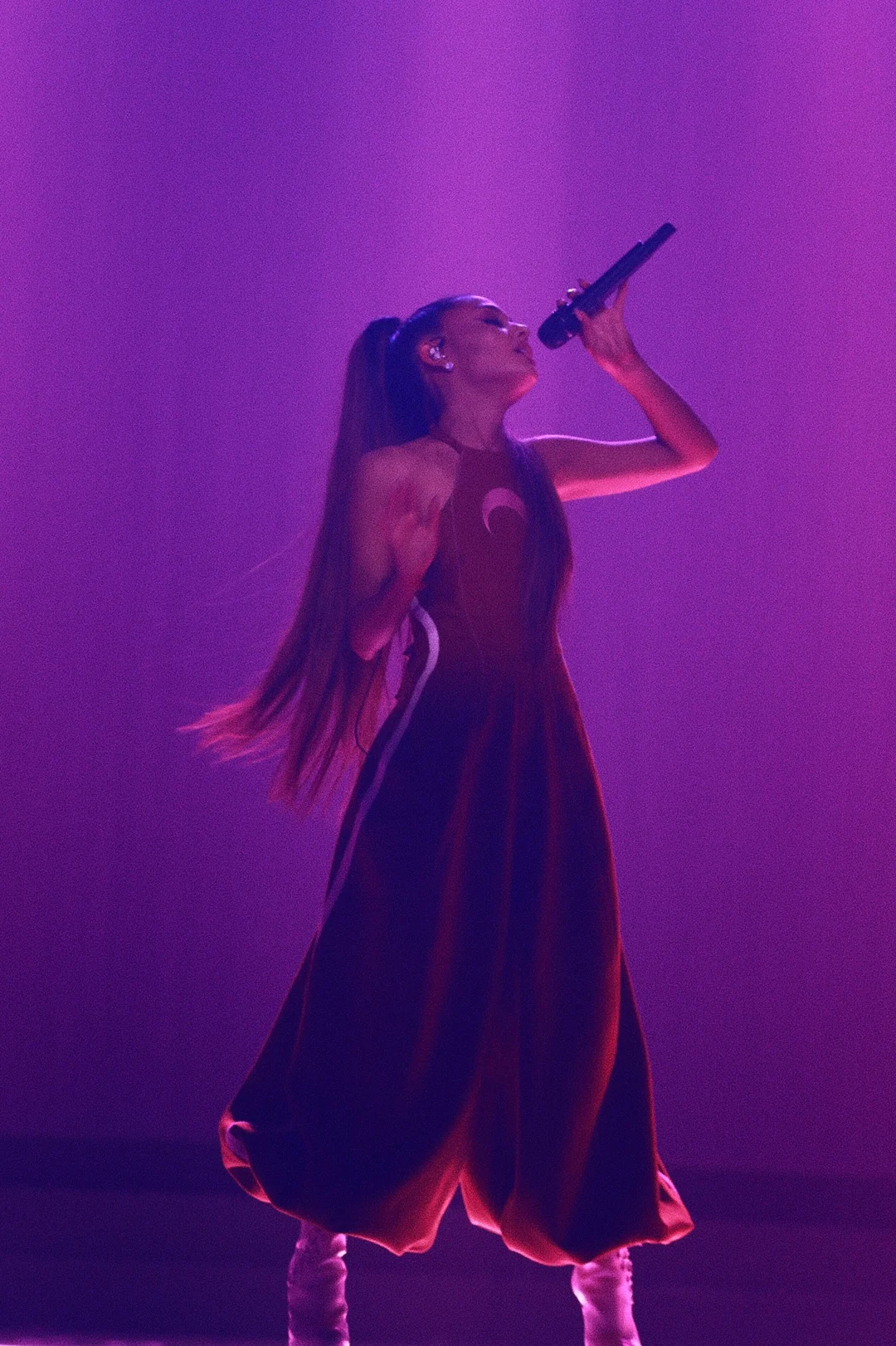 Ariana Grande sings into a microphone on stage, wearing a long dark red dress and high ponytail, bathed in purple stage lighting.