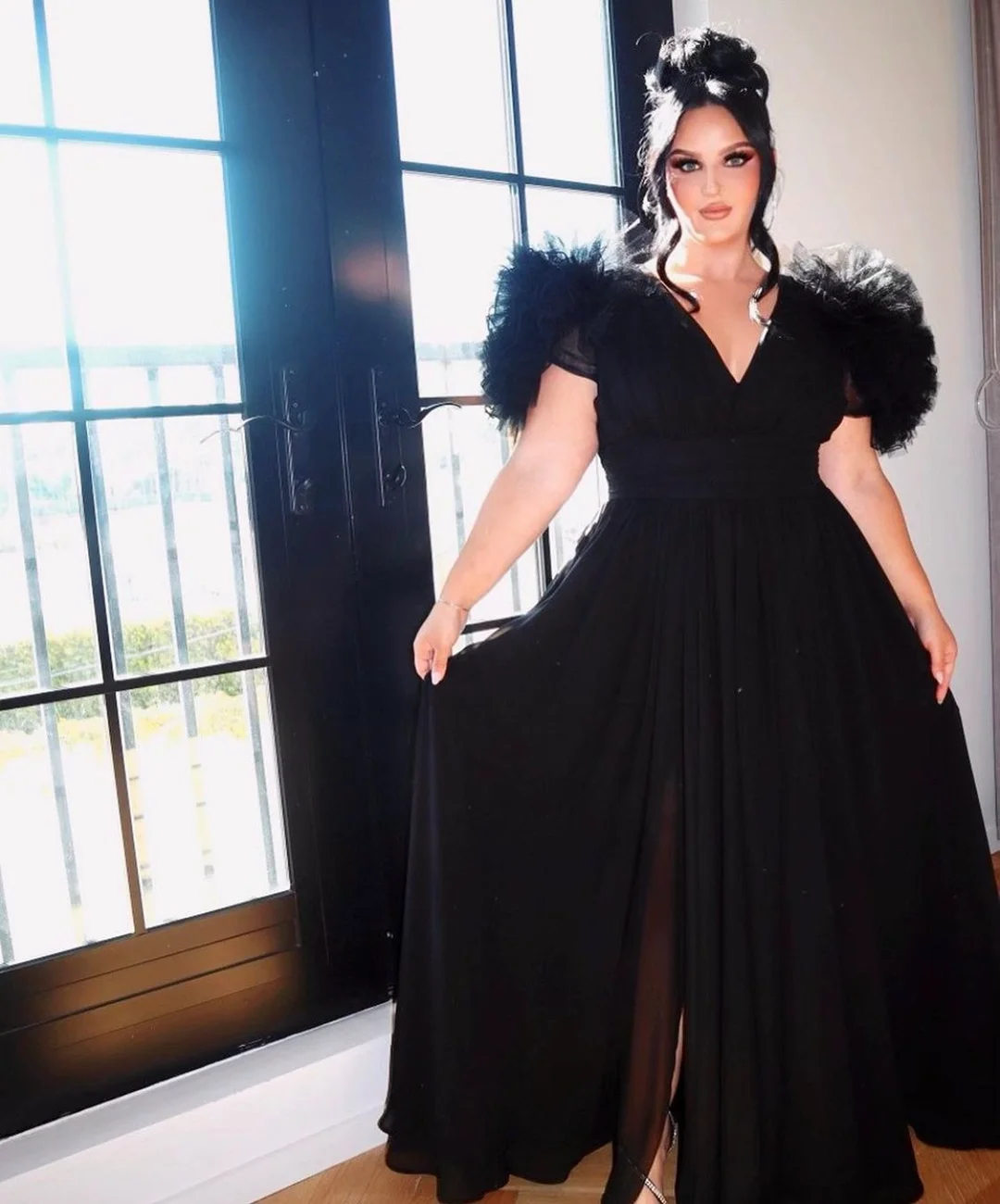 Elegant woman in a long, black gown with tulle sleeves stands beside a window with a view of greenery in the distance.