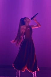 Ariana Grande singing into a microphone on stage under purple lighting, wearing a long red dress with a halter neckline and high ponytail.