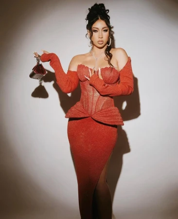 A woman with fair skin poses in a shimmering red evening gown and holding a small purse, standing in front of a light background.