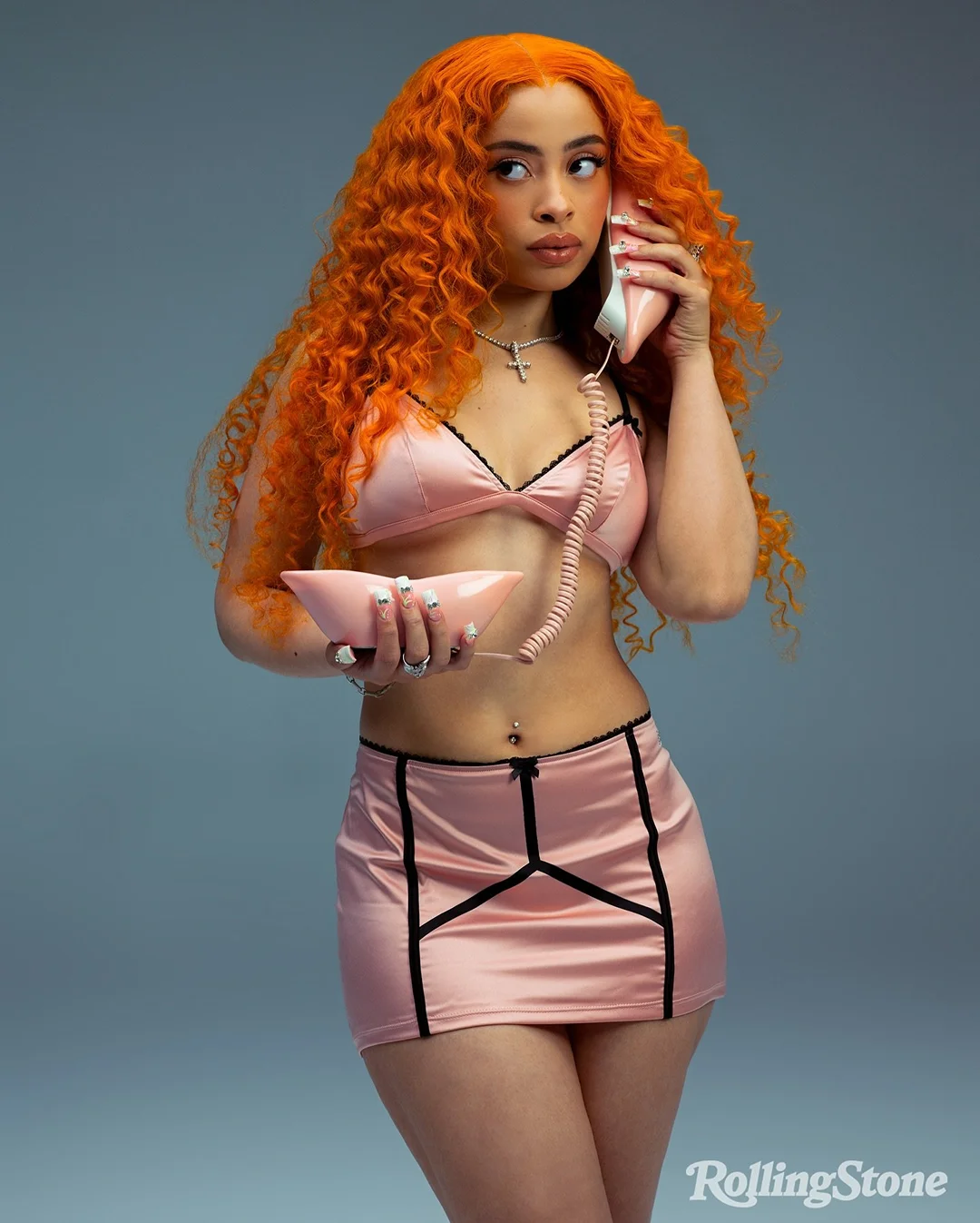 Adult female poses in a pink lingerie set and holds a retro telephone. She has curly orange hair and is in a photo studio.
