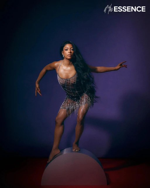 A Black woman strikes a pose on a half-circle podium in a shimmering dress in a studio setting, captured by Essence magazine.
