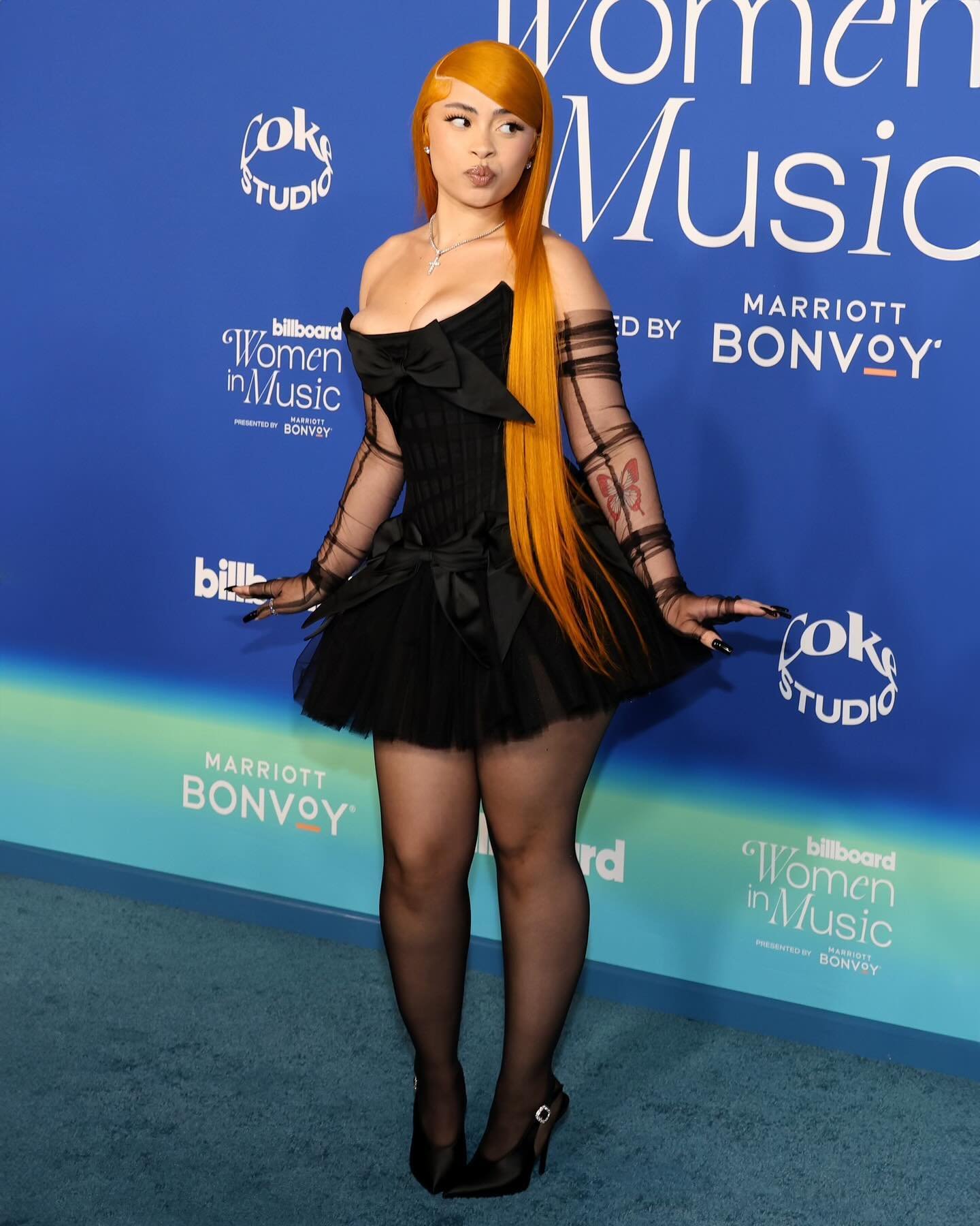 Singer Ice Spice poses in a black dress with a tulle skirt and matching sheer gloves on a blue billboard backdrop at an event.