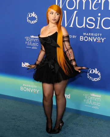 Singer Ice Spice poses in a black dress with a tulle skirt and matching sheer gloves on a blue billboard backdrop at an event.