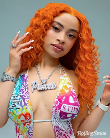 Close-up of Ice Spice with orange curly hair, wearing a colorful halter top, diamond jewelry, and long decorative nails, against a light blue background.