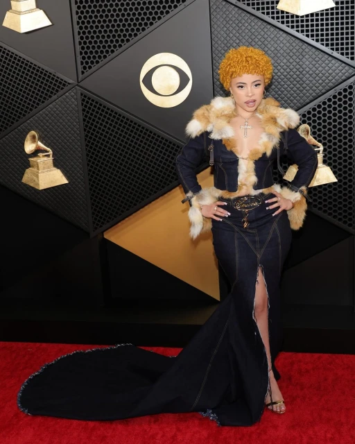 Ice Spice poses on a red carpet in a dark blue denim dress with a fur-trimmed jacket at an awards show, in front of a geometric backdrop.