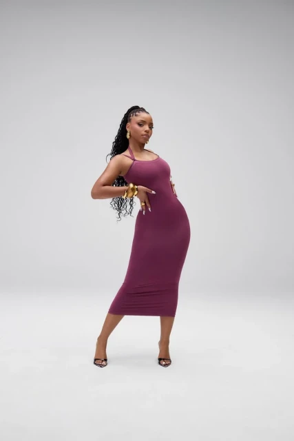 Chloe Bailey poses in a studio wearing a burgundy bodycon dress, gold jewelry, and open-toe heels against a gray backdrop.