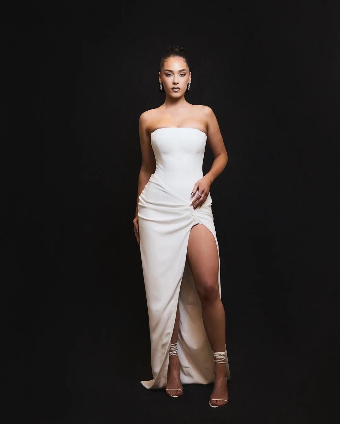 Woman poses in elegant white strapless gown with a thigh-high slit against a dark background in a fashion-style portrait shot.