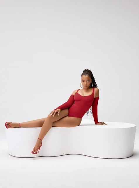 Glamour shot of a Black female model in a red bodysuit and heels, reclining on a white curved platform in a minimalist white studio.