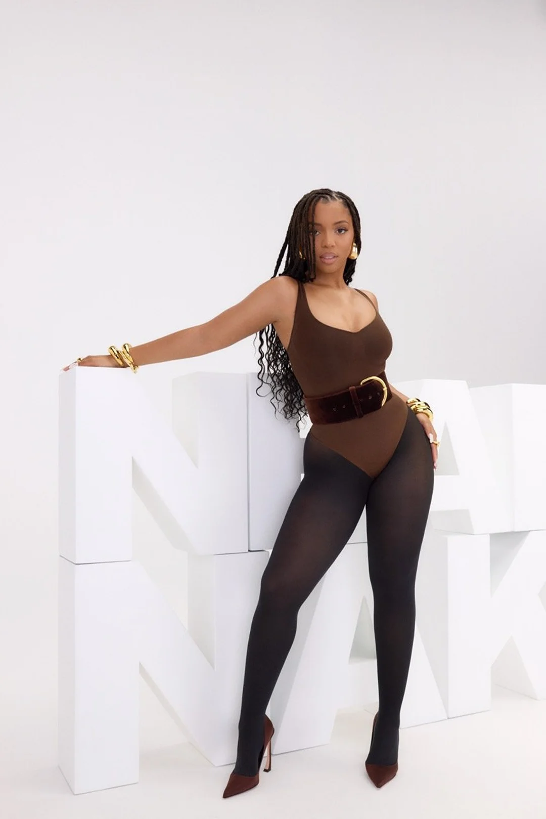 A brunette woman with long braids poses in a brown bodysuit, black tights, and matching heels in a bright studio with block letters.
