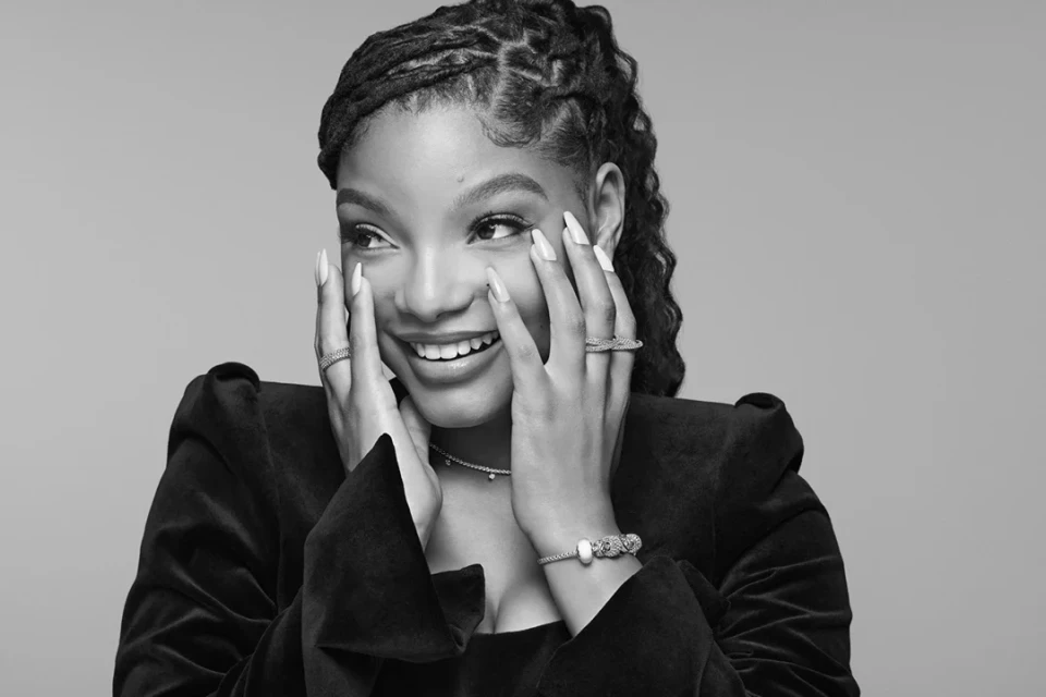 Close-up black and white portrait of smiling adult woman with dreadlocks, wearing a jacket and jewelry against a neutral background.