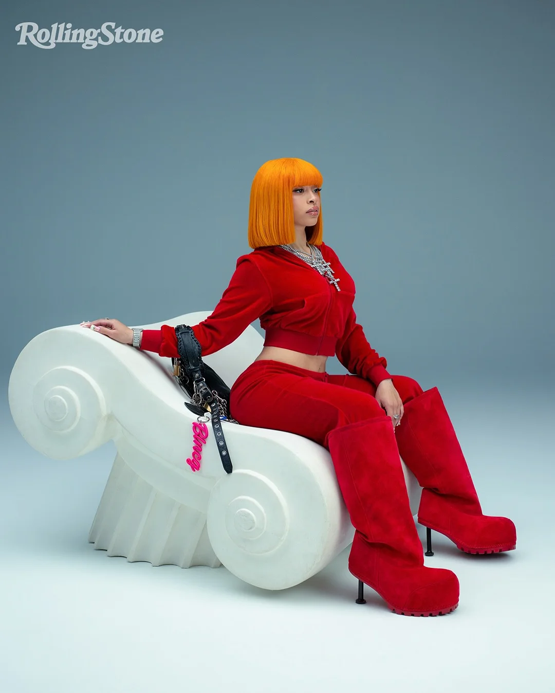 Woman in red velvet tracksuit and orange bob wig sits in a white chair against a grey background for a Rolling Stone magazine shot.
