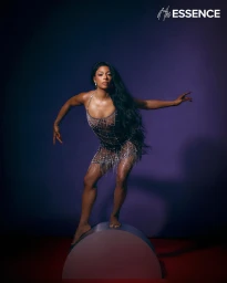 Black woman with long dark hair wearing a crystal-embellished sheer mini dress balancing on a curved metallic prop in a studio.