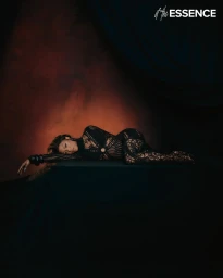 A woman lying on a dark draped surface wearing a black lace jumpsuit, styled by Timothy Luke.