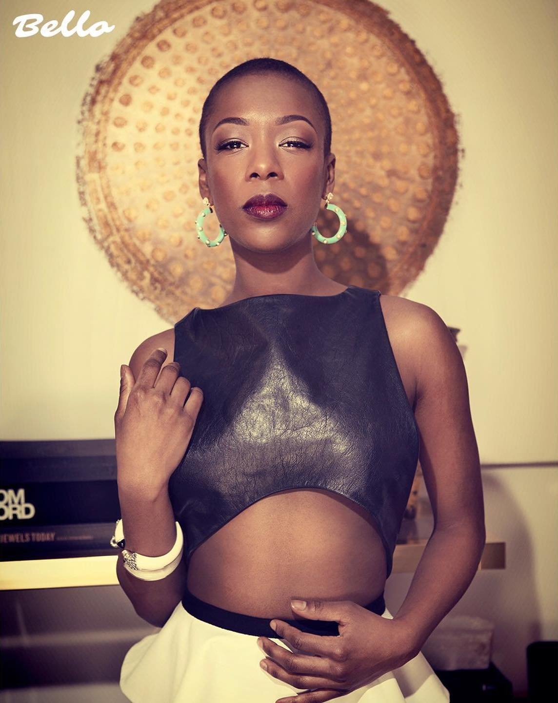 Portrait of a bald Black woman in a black cut-out top with a white skirt, posing in front of a gold-colored decorative wall.