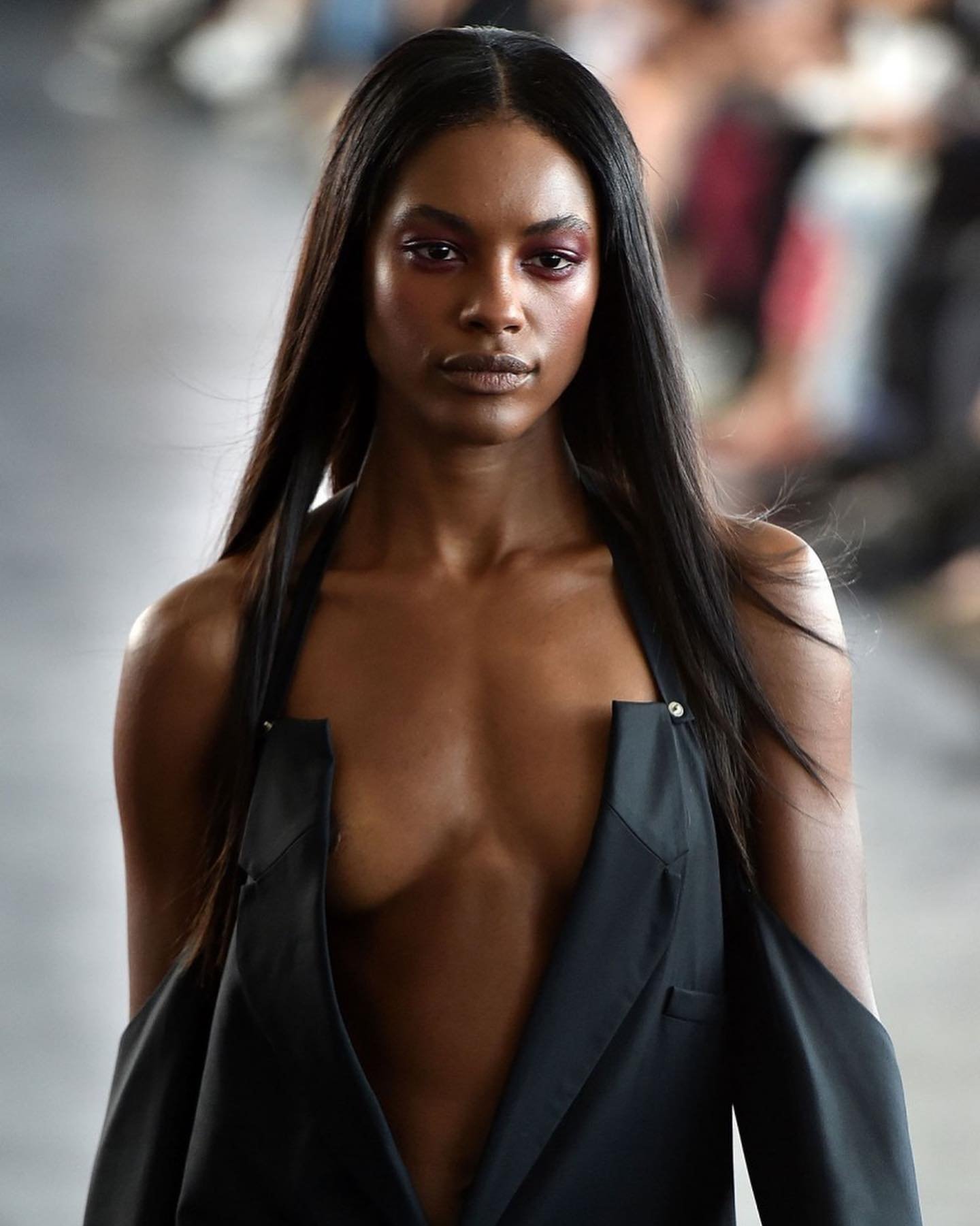 Runway fashion shot of a Black female model wearing a sleeveless black jacket with long, straight hair, posing indoors on a catwalk.