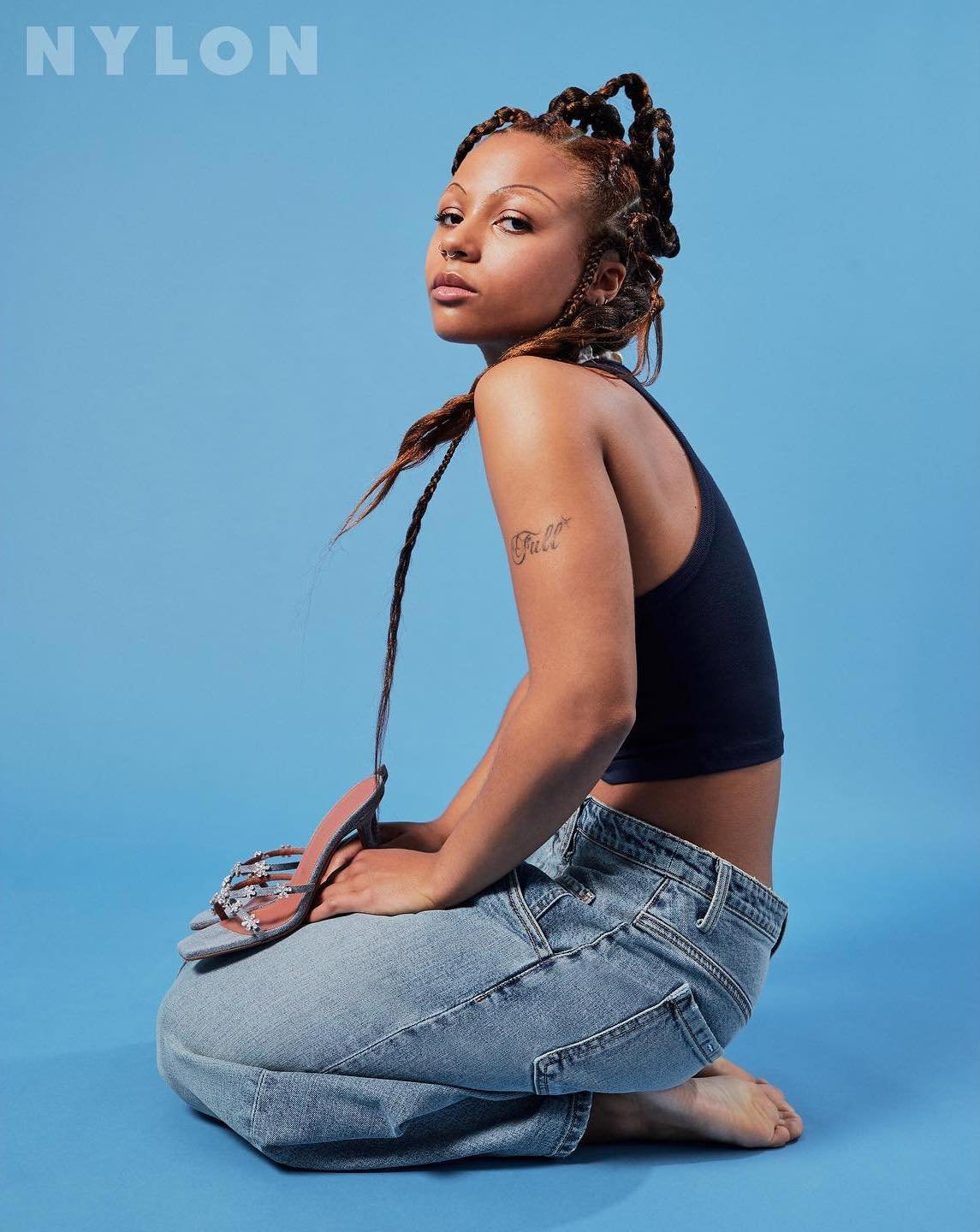 A young adult woman poses kneeling against a light blue backdrop in a tank top and jeans, accessorized with braided hair and sandals.