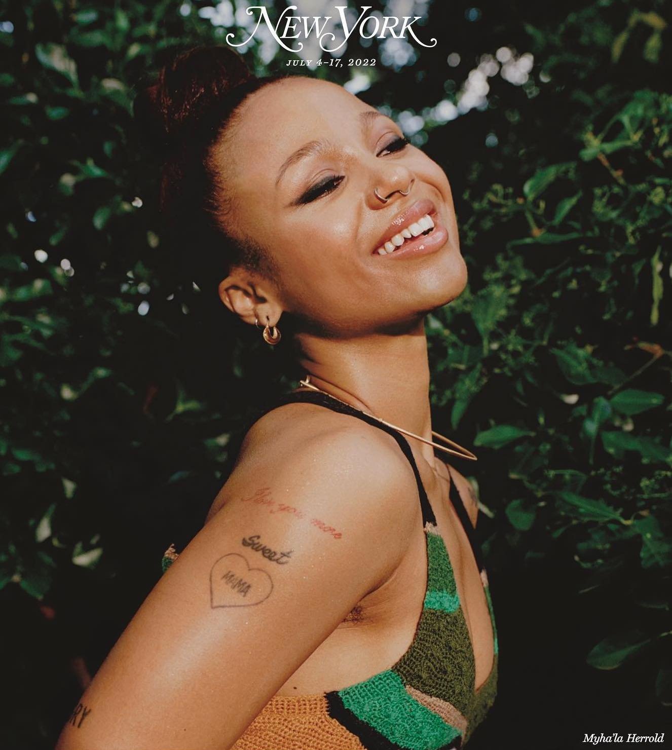 Close-up editorial shot of a smiling woman with a top bun, wearing a knitted top, outside with greenery in the background, on a magazine cover.