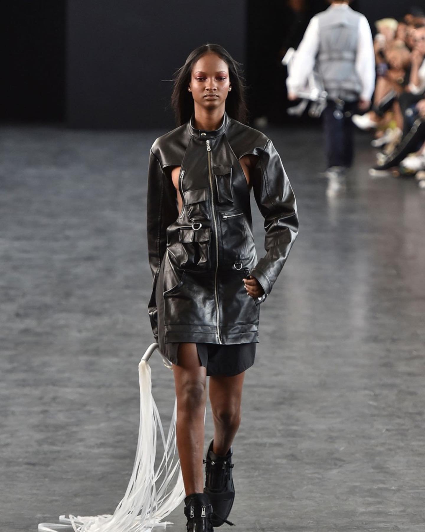A model walks the runway wearing a deconstructed black leather jacket-dress with exposed skin, black platform boots, and trailing white cords.