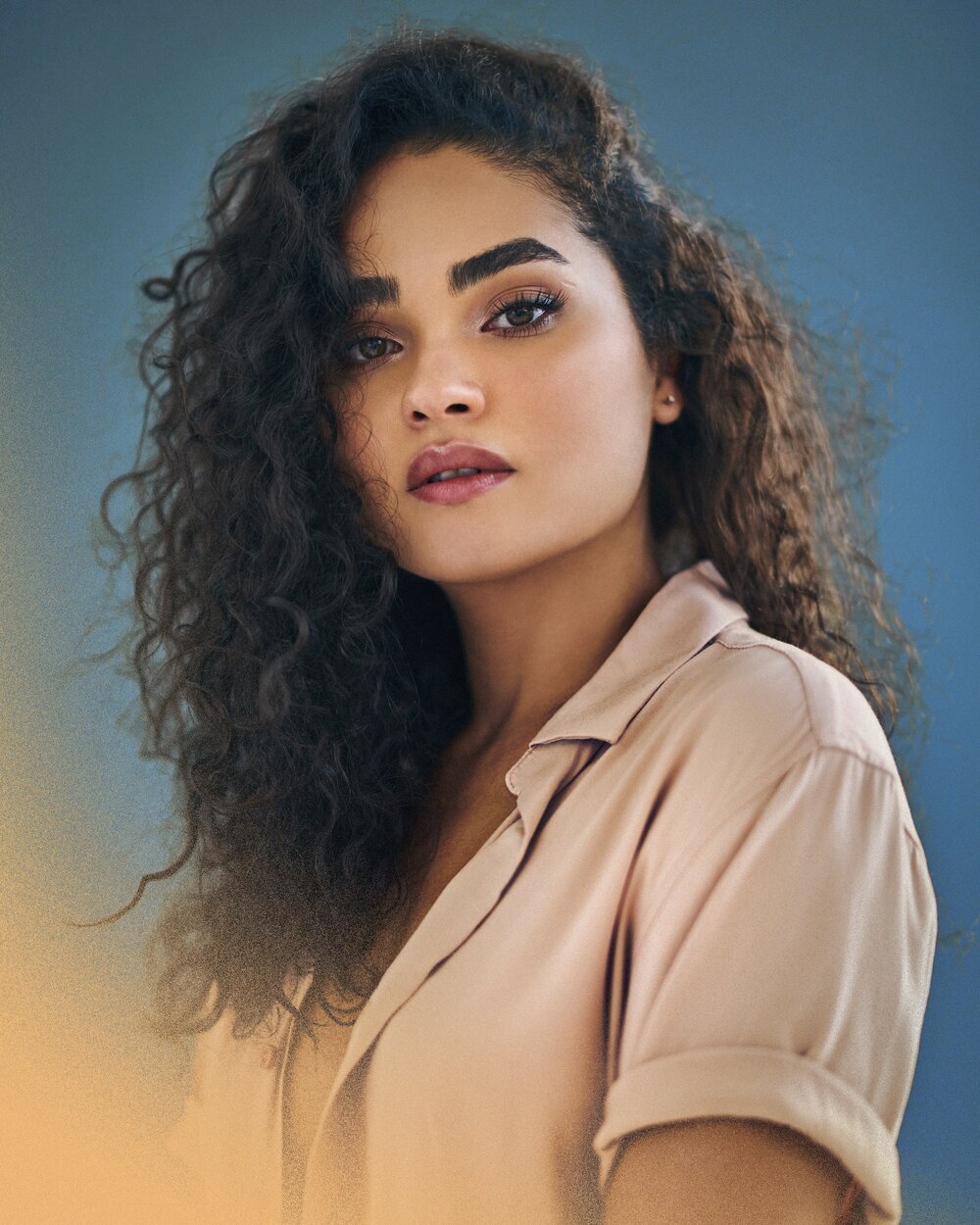 Portrait of a young adult female with curly dark hair wearing a light pink button-down shirt against a blue-toned background in soft lighting.