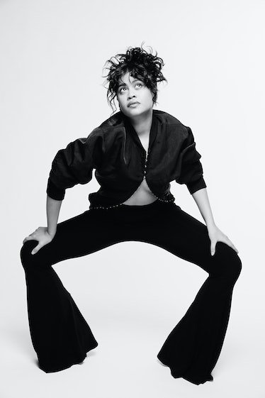 Monochrome portrait of a woman in a black jacket and bell-bottom pants, gazing upward against a light background in a photo studio.