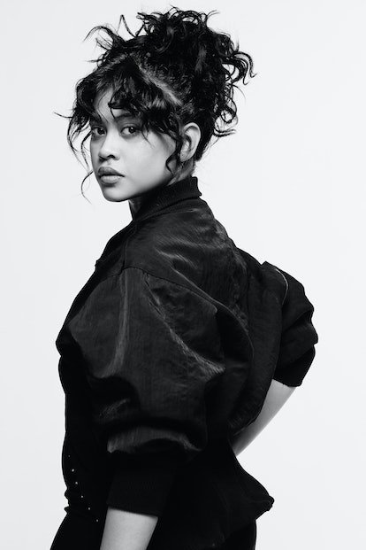Close-up black and white portrait of a woman with curly hair, wearing a jacket, looking over her shoulder in a light studio.